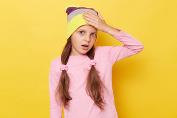 Despair shocked little girl with ponytails wearing pink shirt and beanie hat standing isolated over yellow background having troubles showing facepalm gesture.