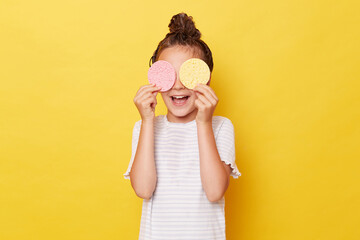 Charming little girl with wet hair wearing casual white T-shirt standing isolated over yellow background posing alone covering eyes with sponges hiding.