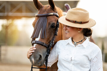 A cute girl in neat clothes and hat is stroking the muzzle of her brown horse as she watches it. The horse is of the Algo-Arabian breed.Concepts of women riding horses, young riders.