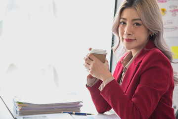 A businesswoman in a red suit holding a cup of coffee to relax in a modern office room before calculating business profit for the company.