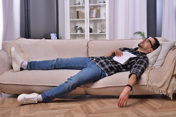 Bookworm's slumber. A man, donning stylish glasses, drifted off to sleep while indulging in a book on a comfortable couch in his inviting living room. Peaceful dreams amid the world of literature.