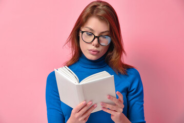 Focused redhead young pretty lady with stylish glasses and blue polo neck reading a book with an...