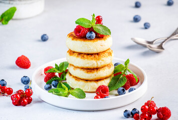 Thick pancakes with cottage cheese with blueberries, raspberries and red currants, decorated with mint on white plate, delicious breakfast. Gray table background