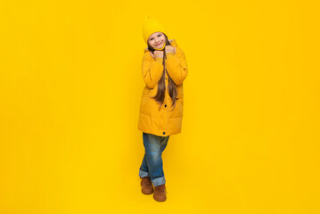 A young girl rejoices at the onset of autumn. Warm clothes for children. A child in an orange down jacket and hat on a yellow isolated background in the studio.