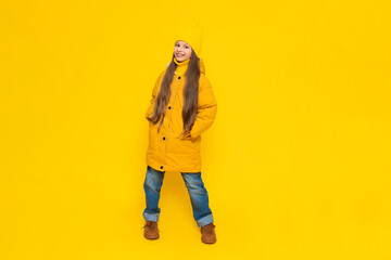 A young girl rejoices at the onset of autumn. Warm clothes for children. A child in an orange down jacket and hat on a yellow isolated background in the studio.