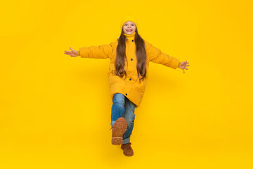 A little girl in full height in an autumn coat, down jackets and hats, rejoices in autumn. The child joyfully raises his leg with his arms outstretched to the sides. Yellow isolated background.