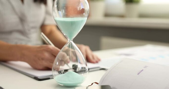 Woman fills out paperwork against the background of hourglass at workplace. Time management and deadline concept