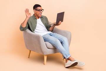 Full length portrait of positive man sit chair arm palm waving hi video call netbook isolated on beige color background