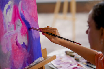 Close-up of a girl artist with a brush applying oil paint to a painting standing on an easel in a studio for painting 