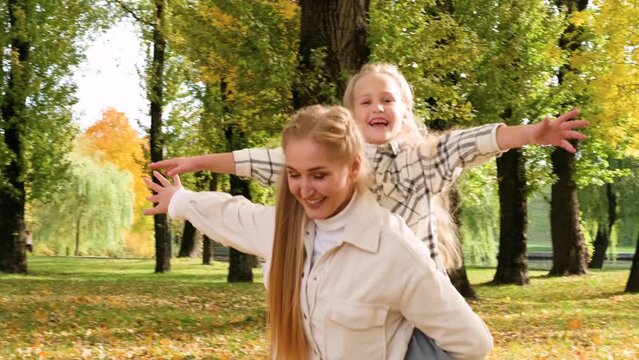 Mother and daughter spend time together in the autumn park. The mother put her daughter on her back and they both depict an airplane. High quality 4k footage