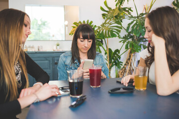 Three young women sitting indoors bar chatting using smartphone drinking soft drink