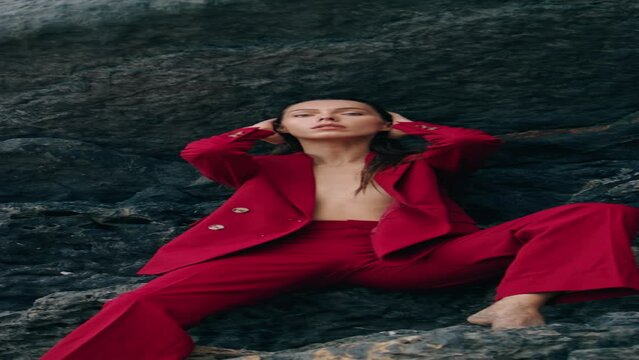 Sexy posh woman posing at cliff in red suit. Girl sitting under rock vertical
