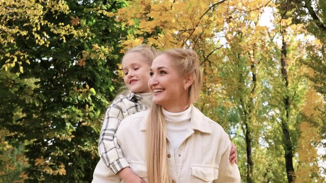 ittle child is having fun with her mother in city park, happy family life, kid girl on her back mom laughs, childish desires play, daughter love hug mom, laughing cheerful people on street glare sun