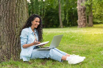 Young Middle Eastern woman sitting in the park and working with laptop, making a notes. Business, education, lifestyle concept. Girl communicates via video call. Language learning, exam preparation