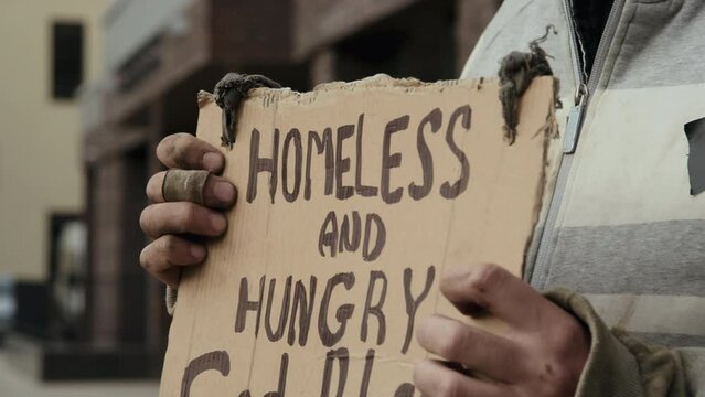 Tilt dow shot of tramp with dirty fingers with sign homeless and hungry standing on street and begging for money