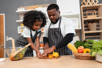 Happy African American kid girl cooking salad with father in kitchen at home	