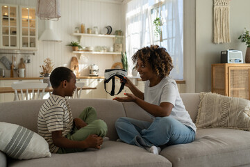 Young African American mother holding VR helmet introducing virtual reality technology to child son while spending leisure time together at home. Mom parent exploring virtual world with kid