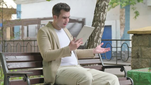 Young Man Upset by Loss on Tablet while Sitting Outdoor on a Bench