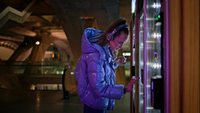 Girl choosing snacks vending machine standing in subway. Woman in headphones.