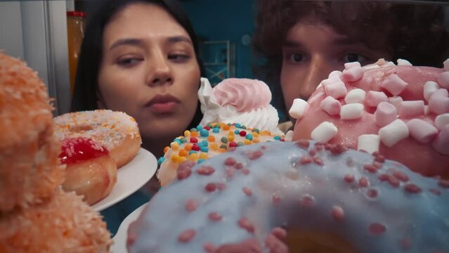 A man and an Asian woman enthusiastically examine desserts in an open refrigerator. A man takes out a cupcake, first gives it to a woman to try, then eats it himself. View from inside the refrigerator