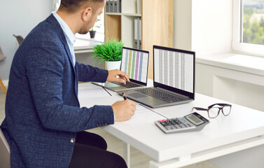 Man in office working with big data and databases using two laptops and excel tables. Office worker making analysis and report with spreadsheets on computer sitting at table near calculator.