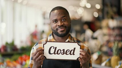 Portrait of a Black-skinned man in a checkered shirt and black apron holding a sign with the Close sign in a supermarket. Video filmed in high quality