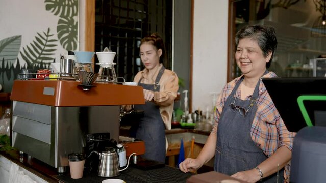 Asian senior woman Serving coffee to customers in front of bar counter In shop,  beautiful Asian barista, owner's daughter, is preparing drinks by the side. Coffee machine page. family small business
