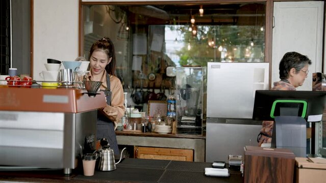 Asian female senior Take coffee from daughter, walk to customers at the table, beautiful Asian barista. Shop owner's daughter Drinks are being prepared on side. coffee maker family small business