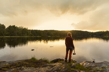 Fototapeta na wymiar A female hiker holding an axe standing in front of a calm lake in the forest watching the sunset.
