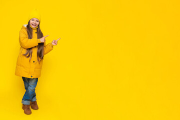 A full-length little girl in a jacket and hat points to the announcement of your advertisement. The child rejoices in autumn. Warm clothes for children. Yellow isolated background. Copy space.