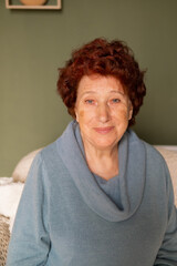 Portrait of an elderly woman with blue eyes and red hair looks at the camera and smiles in the room sitting on the sofa