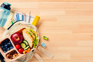 School lunch box with sandwich, vegetables, bottle of water, almonds and fruits and backpack with school supplies on wooden desk table