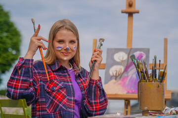 Portrait of a cheerful girl artist with oil paint on her face and brushes in her hands smiling against a city background