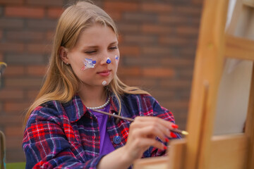 Focused girl artist with paint on her face painting a picture standing on an easel with a...