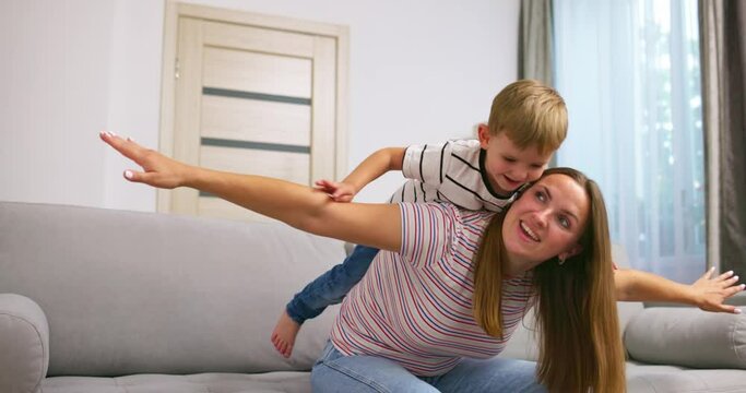 Mother holding on back little son, play together while sitting on couch