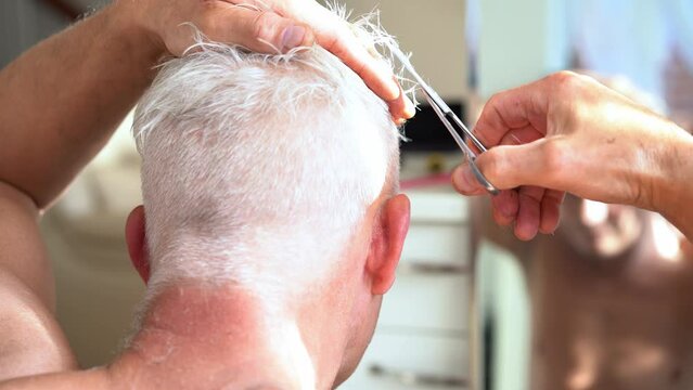 Back view white hair man doing self haircut with scissors and looks in the mirror. Self-care at home in the conditions of global quarantine and closed hairdressers and beauty salons. Close up.