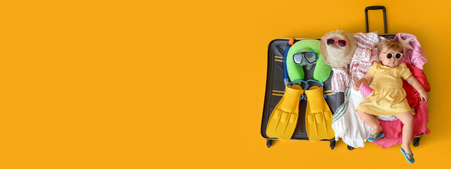Cute baby girl lying in suitcase with belongings on yellow background with space for text, top view