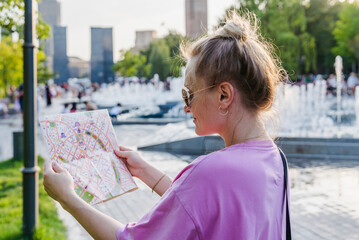 Fototapeta na wymiar Beautiful Smiling Young Woman Tourist Consulting a Paper Map and Looking for Location in a City