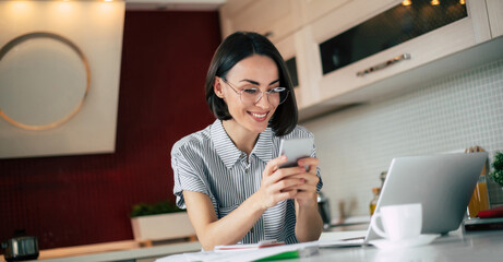 Modern student. Cute and stylish freelance woman in glasses with mobile phone using at laptop and working from home office. Distance learning online education and work.