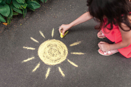A child draws the sun with chalk on the pavement. Selective focus.
A girl draws a yellow sun with chalk on the sidewalk in summer. Summer active holiday concept.