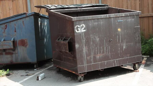 empty garbage dumpster with lid open and another dumpster behind it, surrounded by wood fence in parking lot, full dumpster in shot