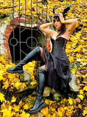 Young woman in a black mask on her head in a black dress and long boots posing near bright orange foliage. Halloween concept.