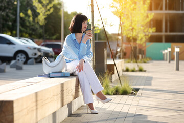 Elegant businesswoman holds paper cup with coffee or tea to go, sitting on wooden bench and relaxing free time outside office building. Female adult worker has lunch break in business area outdoors.
