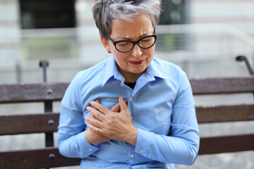 A senior woman suffers from chest pain. She is sitting on a wooden bench in the city. She may have...