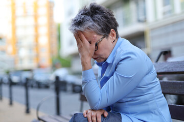 A woman feels sad and tired. She is sitting on a wooden bench on a city street. She may have a headache or a problem.