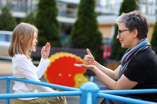A woman and a child communicate with sign language. They are sitting on a blue playground in an outdoor setting. They are mother and daughter or teacher and student. - Powered by Adobe