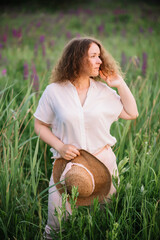 Young woman stands in white shirt in field of purple and pink lupins. Beautiful young woman with curly hair and hat outdoors on a meadow, lupins blossom. Sunset or sunrise, bright evening light