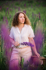 Young woman stands in white shirt in field of purple and pink lupins. Beautiful young woman with curly hair and hat outdoors on a meadow, lupins blossom. Sunset or sunrise, bright evening light