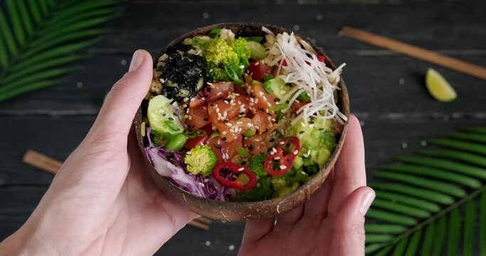 Showing salmon poke bowl, person holding a wooden bowl with raw fish and fresh vegetables, broccoli and edamame beans, high quality video clip, exotic background, palm leaves, 4k footage