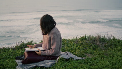 Naklejka na ściany i meble Lonely brunette sitting blanket with book looking gloomy seascape. Romantic girl